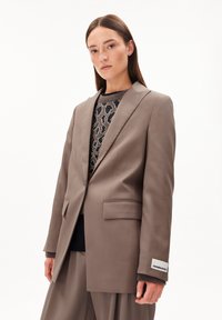 Jonge vrouw in een taupe oversized blazer en bijpassende wijde broek over een leo-print trui, staande tegen een witte achtergrond.