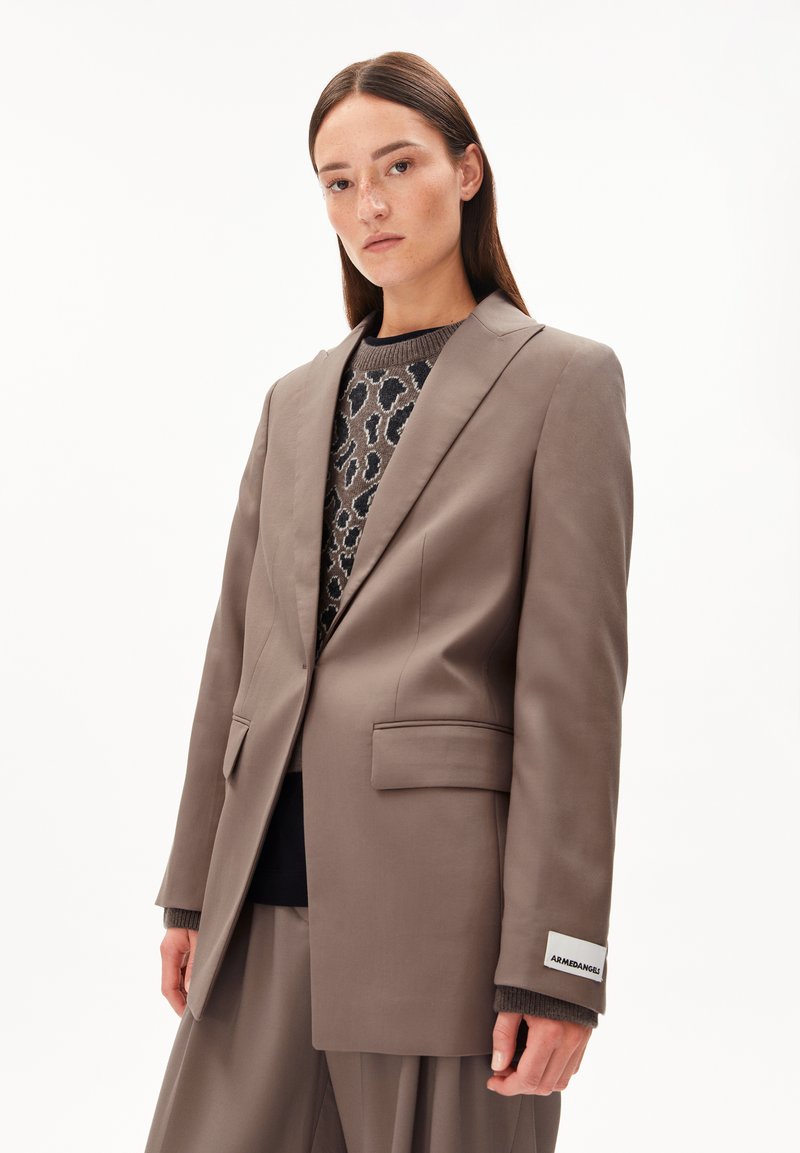 Jonge vrouw in een taupe oversized blazer en bijpassende wijde broek over een leo-print trui, staande tegen een witte achtergrond.