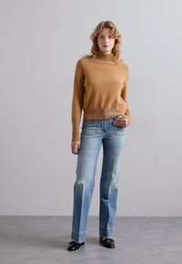 Világosbarna kötött pulóver magas nyakkal, amelyet halványkék farmernadrággal párosítottak, enyhe kopással. Fekete cipőkkel egészíti ki a megjelenést.