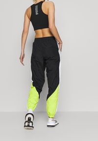 Woman wearing black Puma sports bra, black and neon green track pants, and white sneakers, standing with back facing camera.