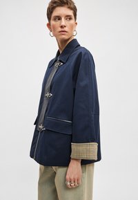 Γαλάζιο μπουφάν με δομημένο σχέδιο, ασημένιες μεταλλικές λεπτομέρειες, φερμουάρ μπροστά και μανσέτες με καρό σχέδιο. Διαθέτει δύο μπροστινές τσέπες.