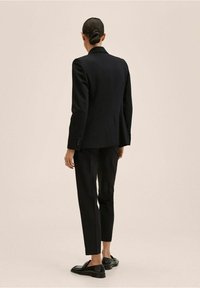 Černé sako na míru s vycpávkami na ramenech, zapínání na jeden knoflík, spárované se slim-fit černými kalhotami a lesklými loafers.