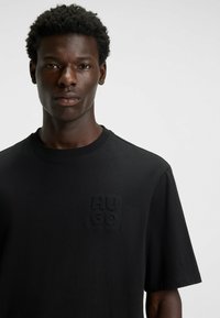 Black cotton T-shirt featuring a crew neckline and short sleeves. The front displays an embossed "HUGO" logo on the left chest area.