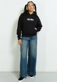 Czarna bluza z kapturem z grafiką, z przednią kieszenią i ściągaczem, w połączeniu z jasnoniebieskimi spodniami dżinsowymi w szerokim kroju i czarnymi butami wsuwanymi.