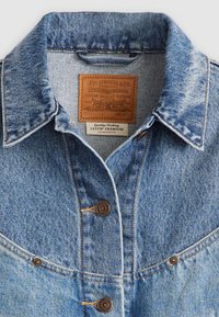 Svetlo modra denim jakna z ostrim ovratnikom, rjavimi gumbi in usnjeno etiketo znamke z reliefnimi podrobnostmi na prsnem predelu.