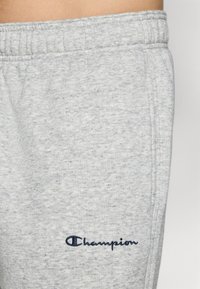 Lichtgrijze joggingsbroek van zacht, gestructureerd materiaal met een elastische tailleband en een blauw geborduurd "Champion" logo aan de linkerkant.