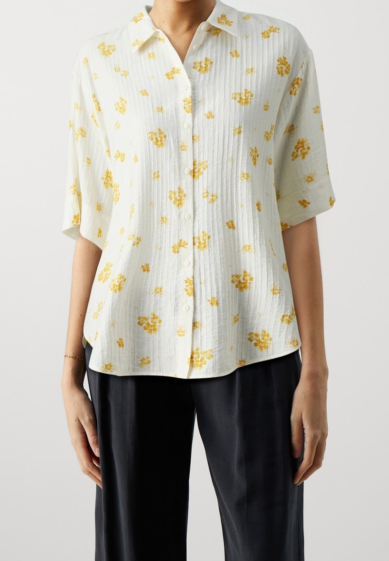 Blouse blanche à manches courtes boutonnée avec un imprimé floral jaune, associée à un pantalon noir ample sur une personne debout.