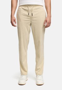 Beige corduroy pants with an elastic waistband and drawstring. Features a straight fit and subtly textured surface, paired with white sneakers.