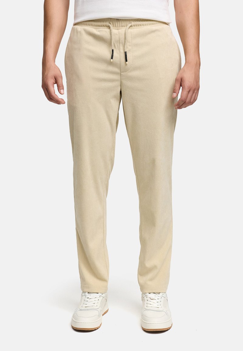 Beige corduroy pants with an elastic waistband and drawstring. Features a straight fit and subtly textured surface, paired with white sneakers.