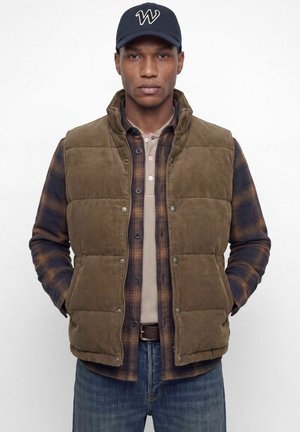 Un homme porte un gilet matelassé marron par-dessus une chemise à carreaux et un haut beige, avec un jean et une casquette bleu marine, sur un fond blanc uni.