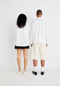 To personer står rygg mot rygg. Den ene har på seg en lang hvit skjorte og et svart plisséskjørt, den andre har på seg en hvit skjorte og beige shorts.