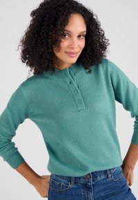 Türkiser Strickpullover mit Henley-Ausschnitt, drei Knöpfen und gerippten Bündchen. Weiche Textur, mittlere Passform, kombiniert mit Denim-Jeans.