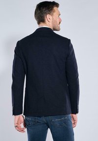 Navy pinstripe blazer with slim fit, notched lapels, single vent back, and two buttons. Worn over blue jeans, showing textured material.