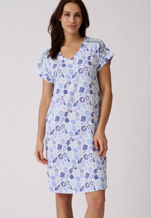 Mujer con vestido blanco hasta la rodilla con estampados azules de conchas marinas y estrellas de mar, mangas cortas y escote en V, de pie frente a un fondo liso.