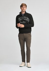 Svart zip-up sweatshirt med texten "Polo Club", i kombination med bruna byxor och ljusa sneakers. Modellen står med händerna hopknäppta.