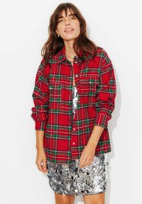 Chemise en flanelle à carreaux rouges avec des accents verts, dotée d'une fermeture à boutons et de deux poches avant, portée sur une mini-jupe argentée à sequins.