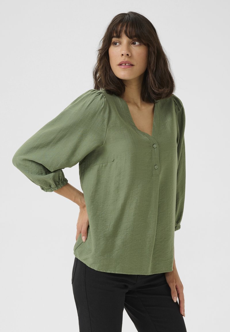 Groene, gestructureerde blouse met een V-hals, 3/4 pofmouwen, en twee knopen aan de voorkant. Gepaard met zwarte broek met zijzakken.