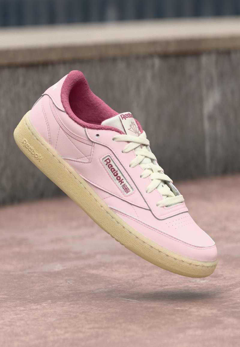 Sapatilhas cor-de-rosa claro com parte superior em couro liso, atacadores em creme e sola de borracha bege. Apresenta um forro interior rosa e detalhes com o logo da Reebok.