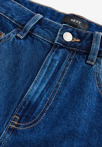 Jean en denim bleu foncé avec une fermeture à bouton métallique, des coutures visibles, et une étiquette "NEXT" sur la ceinture.