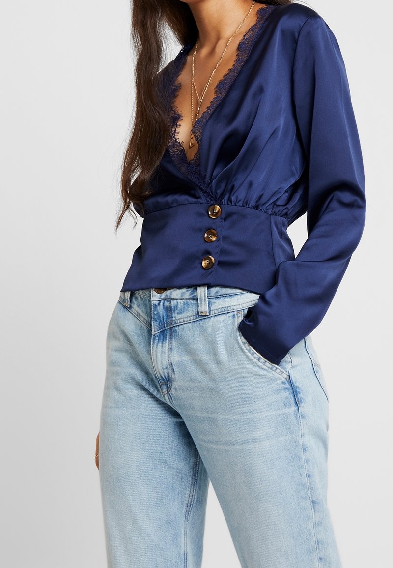 Femme portant un chemisier en satin bleu marine à manches longues avec bordure en dentelle et trois boutons, associé à un jean taille haute bleu clair, main dans la poche.