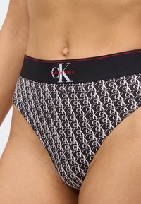 Μαύρο εσώρουχο στρινγκ με επαναλαμβανόμενο μοτίβο λευκού λογότυπου "CK". Η μέση έχει μαύρη ταινία με την επιγραφή "Calvin Klein" και κόκκινες λεπτομέρειες.