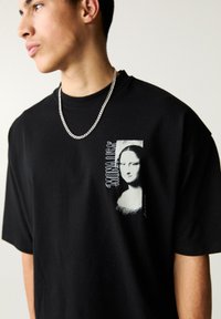 Schwarzes Baumwoll-T-Shirt mit einem Grafikdesign der Mona Lisa, mit weißem Text und Designelementen, locker geschnitten und mit kurzen Ärmeln.
