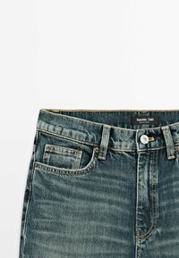 Taille de jean en denim bleu avec bouton frontal, passants de ceinture et étiquette Massimo Dutti visible à l'intérieur de la ceinture.