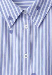 Chemise rayée verticalement bleu et blanc avec col boutonné, boutons blancs et petits cœurs bleus brodés près du col et de la poitrine.