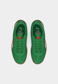 Ζευγάρι πράσινα αθλητικά παπούτσια Puma με κόκκινο λογότυπο, λευκές λεπτομέρειες και καστανές σόλες, από πάνω σε λευκό φόντο.