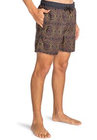 Hombre con pantalones de baño negros con patrones geométricos circulares en amarillo y marrón, de pie descalzo con la mano en el bolsillo.