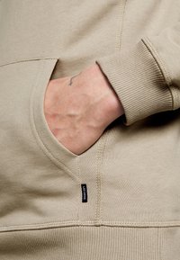 Main avec petit tatouage de croix insérée dans la poche d'un sweat beige à poignets côtelés avec étiquette Les Deux visible.