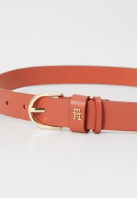 Cintura in pelle marrone chiaro con una fibbia in metallo dorato, dotata di una tracolla con marchio e più fori per la regolazione della misura. Texture liscia in tutto.