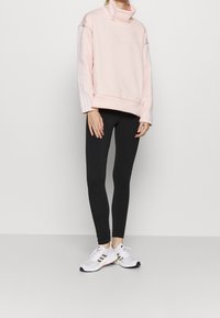 Pull-over rose clair avec col montant et cordon blanc, orné de trois bandes blanches sur les manches ; associé à des leggings noirs et des baskets blanches.
