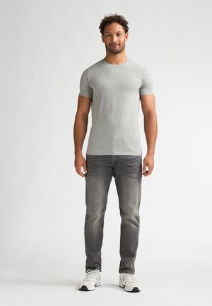 T-shirt gris à manches courtes associé à un jean gris foncé et des chaussures de sport blanches. L'homme est debout, face à l'appareil, sur un fond neutre.