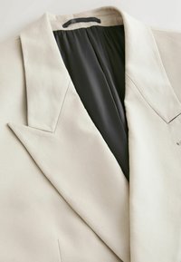 Blazer beige clair avec un large revers cranté et une doublure intérieure noire, montré en gros plan.