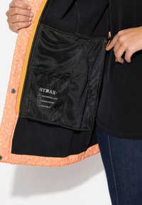 Chaqueta naranja ligera y repelente al agua con patrones de gotas blancos; cuenta con un forro interior negro con una etiqueta que indica atributos de resistencia al agua y transpirabilidad.