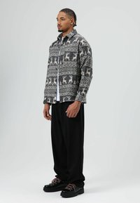 Chemise boutonnée en tissu gris et crème à motifs de rennes, associée à un pantalon noir ample et des chaussures noires épaisses.