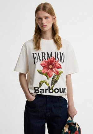 Νεαρή γυναίκα με μακριά μαλλιά που φοράει μια λευκή μπλούζα με ένα μεγάλο κόκκινο λουλούδι και το κείμενο «FARM RIO Barbour», κρατώντας μια φλοράλ καπέλο.