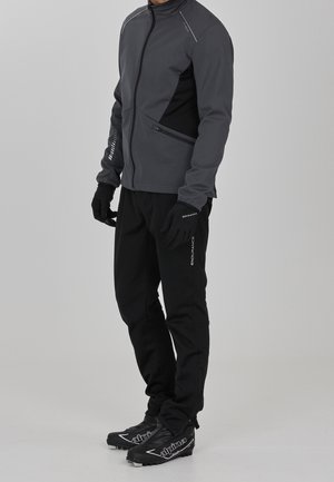 Mann trägt graue Jacke mit Reißverschluss, schwarze Handschuhe, schwarze Hose mit dem Schriftzug "ENDURANCE" und schwarze "Alpine" Langlaufskischuhe, steht vor grauem Hintergrund.