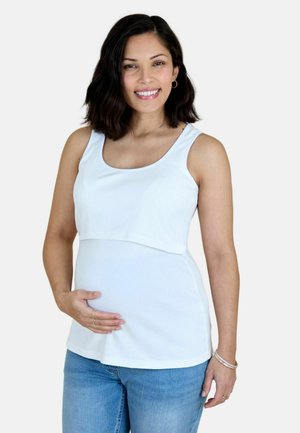 Lachende zwangere vrouw met een wit mouwloos topje en een blauwe spijkerbroek, die haar buik voorzichtig met één hand vasthoudt.