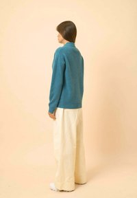 Turkos stickad tröja med krage, ihoptagen med oversize vita byxor. Slät textur, avslappnad passform, visad bakifrån mot en ljus bakgrund.