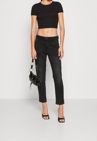 Kvinna iförd svart kortärmad t-shirt, högmidjade svarta jeans, svarta öppna klackar, hållande en svart handväska med metallringdetalj.