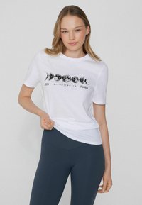 Camiseta de algodón blanca con un gráfico de fases de la luna en negro. Mangas cortas, cuello redondo, combinada con leggings oscuros. Diseño sencillo y casual.