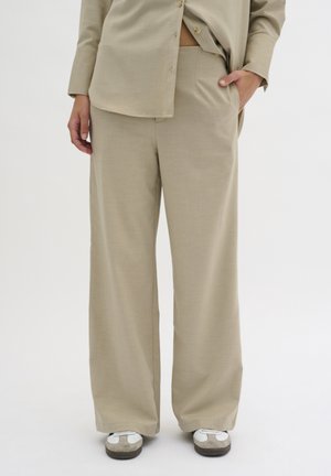 Persoon draagt beige wijde broek en bijpassend beige overhemd met knopen, staat met één hand in de zak, witte en grijze sneakers zichtbaar.
