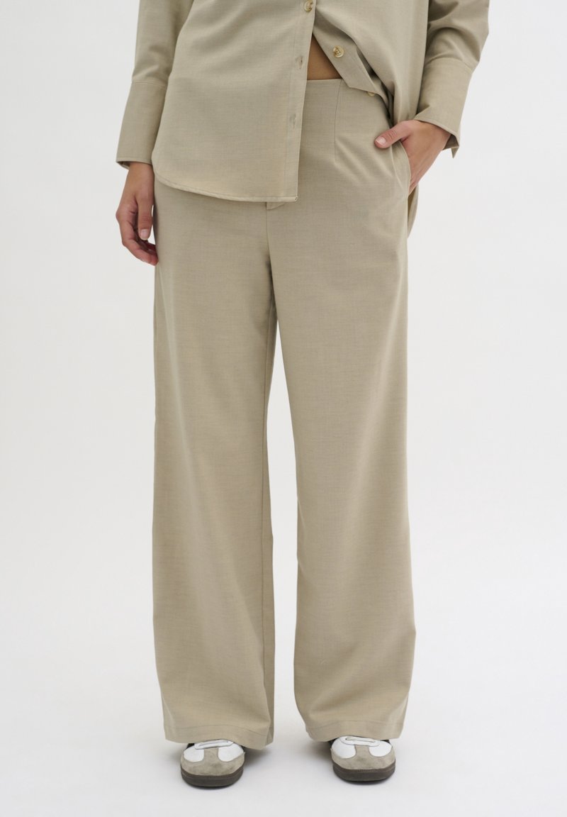 Personne portant un pantalon beige à jambes larges et une chemise beige assortie boutonnée, debout avec une main dans la poche, baskets blanches et grises visibles.