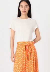 Cremfarbener T-Shirt mit kurzen Ärmeln, kombiniert mit einem knielangen orangefarbenen Rock mit weißen Blättermustern und einem gebundenen Bund. Glatter, leichter Stoff.