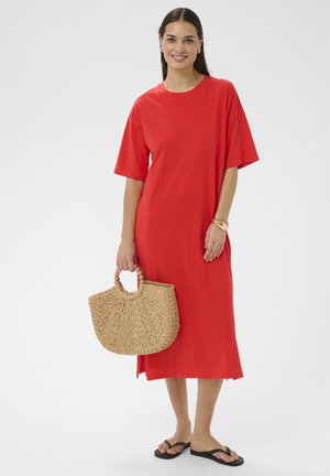 Kvinde i en løs rød midikjole med en flettet stråhåndtaske og sorte sandaler, der smiler foran en ensartet lys baggrund.