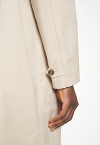 Dunkelhäutige Hand ruht neben dem Ärmel eines beigen Trenchcoats mit sichtbarem, geknöpftem Bundriemen.