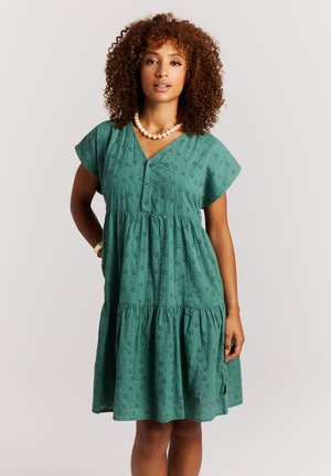 Vrouw met krullend haar, gekleed in een groene jurk met oogjes, parelketting en polshorloge, staand tegen een eenvoudige lichte achtergrond.