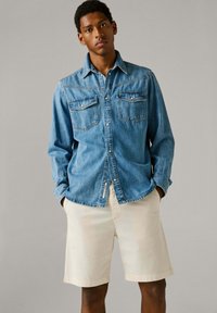 Νέος άντρας που φοράει ανοιχτό μπλε τζιν πουκάμισο με μακριά μανίκια και κρεμ σορτς μέχρι το γόνατο, στέκεται με τα χέρια στις τσέπες.
